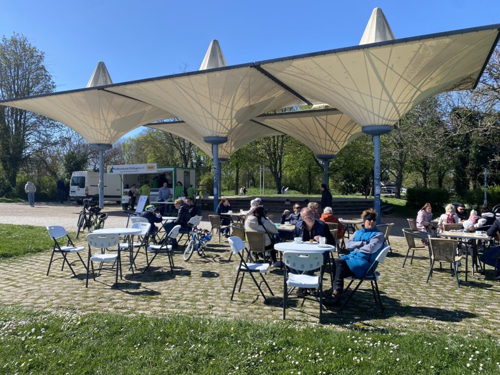 Mehrere Menschen sitzen an Tischen mit Stühlen im Horbachpark beim Kafeehäusle im Freien an einem sonnigen Tag